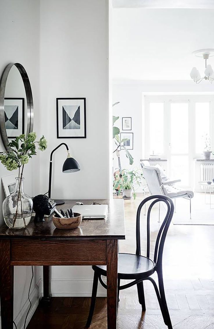 A serene and stylish workspace featuring a vintage wooden desk with a marble top, holding a glass vase with fresh greenery, a black table lamp, and a bowl of art supplies. A traditional bentwood chair complements the desk, positioned against a white interior wall adorned with framed abstract art and a round mirror, with a view into another room showing a comfortable reading chair and indoor plants.
