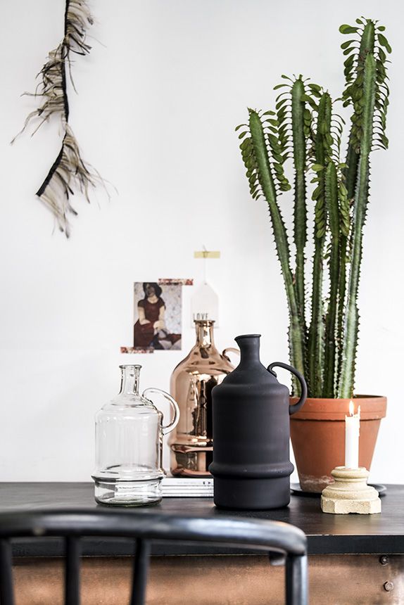 Bohemian chic decor on a black table featuring a matte black jug, transparent glass bottle, a lit candle on a stone holder, and a terracotta pot with a tall cactus. In the background, wall art with feathers and a framed picture add a touch of artistic flair, while a copper distiller enhances the eclectic aesthetic.