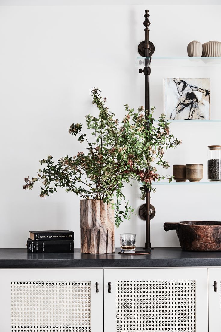 Modern minimalist interior design with a natural wood vase filled with lush greenery on a dark countertop, complemented by a classic 'Great Expectations' book stack, a glass of whiskey, and a rustic wooden bowl. In the background, a sleek lamp stands next to an abstract monochrome artwork and ceramic vases on a floating shelf.