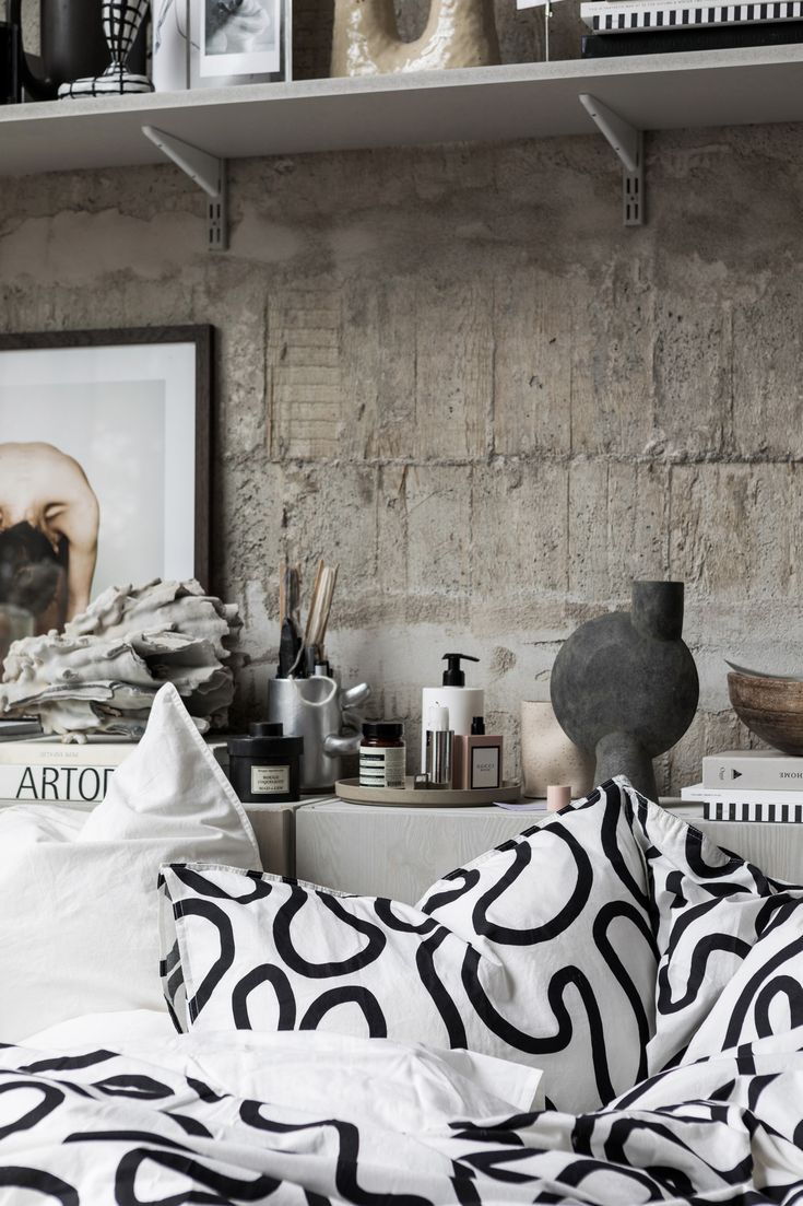 An industrial-style bedroom corner with a prominent raw concrete wall. On the bed are striking black and white graphic-patterned textiles. A bedside area features an array of items including a large rustic vase, a wooden bowl, assorted candles, skincare products, and a stack of books with 'ART' visible on the spine. Above, white shelves hold minimalistic decor and framed artwork, contributing to the room's modern artistic flair.