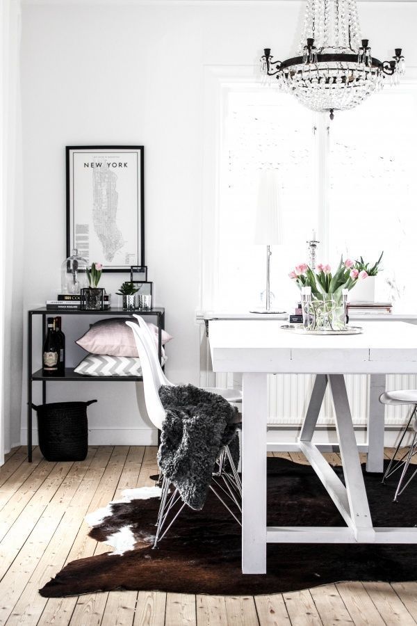Elegant dining nook with a modern-meets-classic flair, showcasing a sparkling crystal chandelier above a white table adorned with fresh pink tulips. The room features a plush sheepskin-draped chair over a dark cowhide rug, with a sleek black console table against the wall holding chic decor and a framed New York map print. Light wood floors and a bright window enhance the room's airy ambiance.