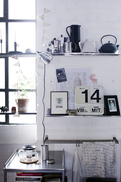 An artistic kitchen corner with a modern yet homey vibe, featuring a stainless-steel countertop with coffee-making essentials including a kettle, mugs, and a French press. The wall above is adorned with a variety of framed prints and personal memorabilia, anchored by a large '14' graphic, against a stark white-painted brick backdrop. The arrangement is illuminated by natural light streaming through a black-framed window, complemented by the warm glow of a metallic desk lamp.