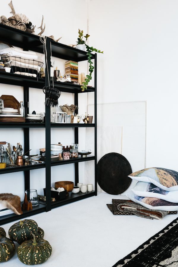 A cozy nook with a black open shelving unit displaying an eclectic mix of items including books, blankets, and kitchenware. A green vine dangles elegantly from the top shelf, adding a touch of nature. On the floor, decorative gourds, a variety of patterned pillows, and a textured rug create a welcoming, bohemian vibe against the stark white wall.