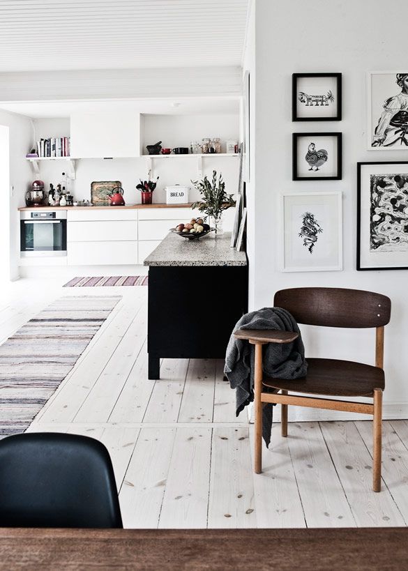 Minimalist Nordic kitchen space highlighted by white walls and light wood flooring. A central black kitchen island with a speckled countertop is adorned with a simple vase and fruit, facing a wall of sleek white cabinetry and shelves decorated with assorted kitchenware and framed artwork. A wooden chair with a throw blanket and a striped rug add warmth and texture to the scene.