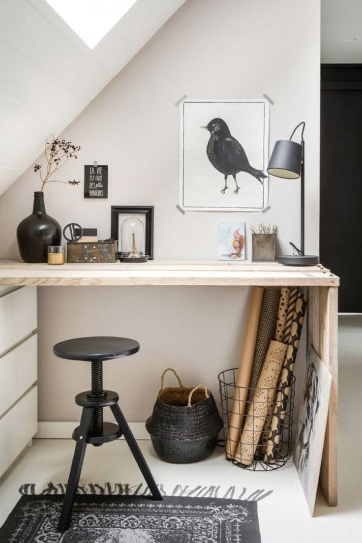 A stylishly minimalist attic workspace featuring a sleek black stool, a wooden desk adorned with eclectic decor including a large framed illustration of a crow, various small framed art pieces, a dark vase with dried branches, and rolls of decorative paper. A black wall lamp adds a modern touch to the serene space.