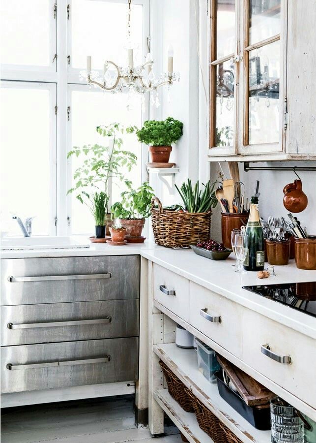 A cozy corner of a kitchen bathed in natural light from large windows, highlighting a sophisticated blend of rustic charm and industrial chic. The workspace is adorned with a collection of potted herbs on the windowsill and a variety of kitchen utensils and copper mugs arranged neatly on the counter. A white cupboard with silver handles provides storage, juxtaposed with a sleek, metal drawer unit. A crystal chandelier adds a touch of elegance above, while wicker baskets below hint at practical storage solutions in this harmoniously styled kitchen scene.