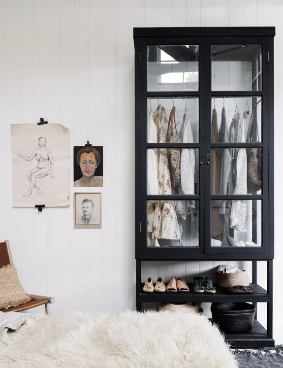 A chic and artistic bedroom corner with a white shaggy rug on the floor. To the left stands a wooden chair with a woven seat, and the wall is adorned with a series of vintage-style drawings and portraits, adding a touch of personal history. A tall black cabinet with glass doors reveals an organized display of clothing and accessories, with shoes neatly lined on the bottom and decorative bowls on a shelf. The white paneled wall enhances the contrast and contributes to the space's eclectic elegance.