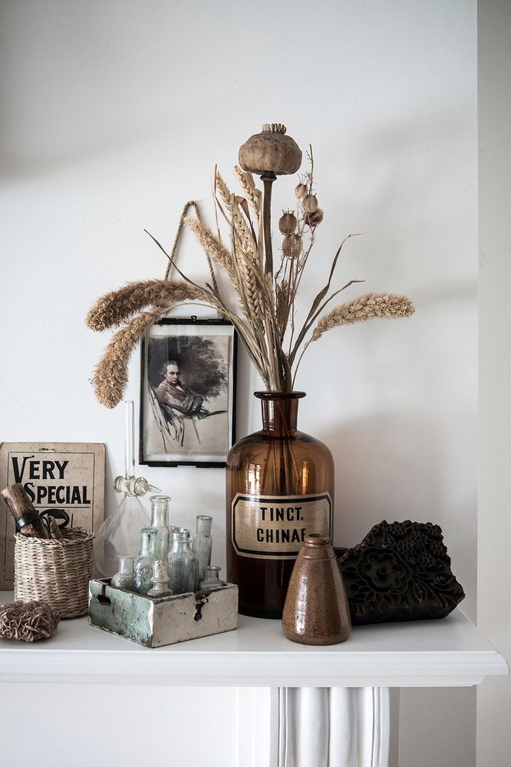 A rustic home decor display with dried botanicals in a vintage brown pharmacy bottle marked 'TINCT. CHINAF,' alongside an old monochrome portrait, a collection of small glass vials, and a woven basket with a sign reading 'VERY SPECIAL' on a white shelf.