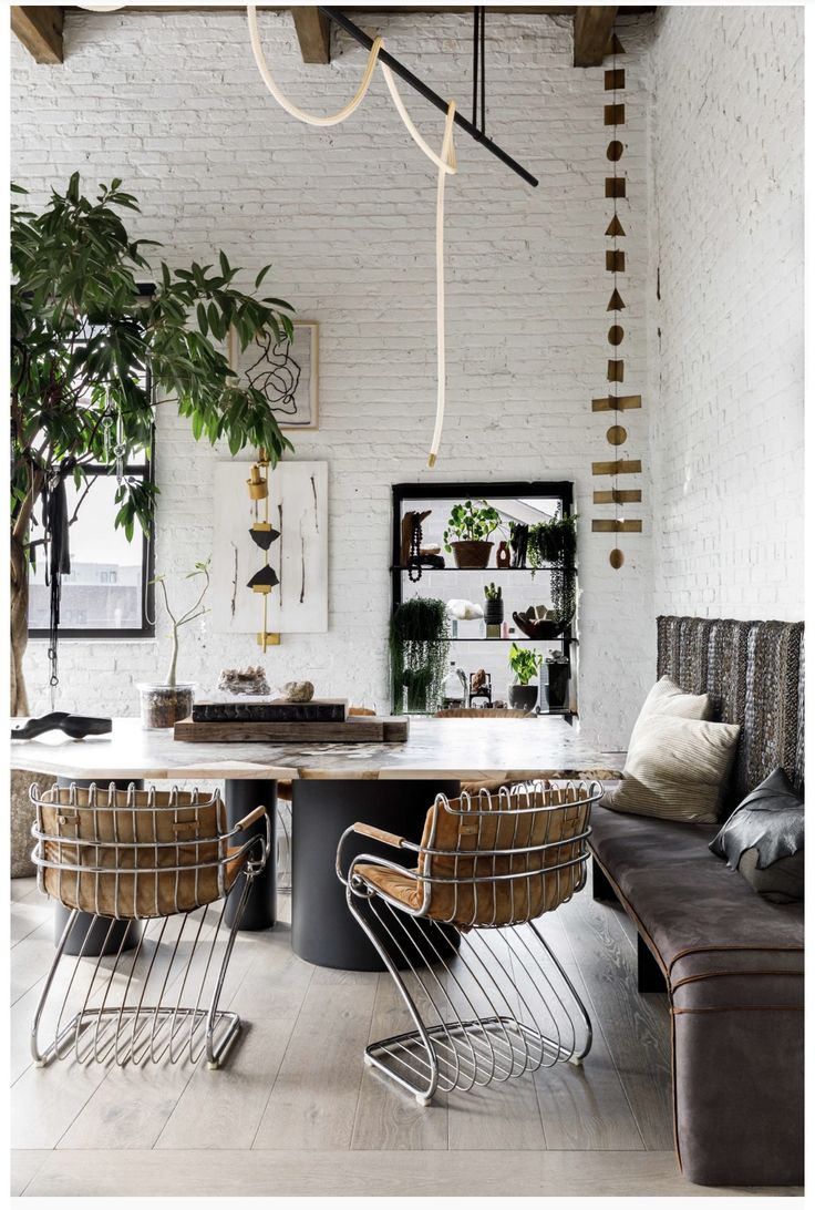 An elegantly styled living space featuring a whitewashed brick wall, with a modern black centerpiece table surrounded by unique wire-frame chairs with leather cushions. A corner sofa sits beside a windowed wall adorned with hanging plants, reflecting a cozy and inviting atmosphere.