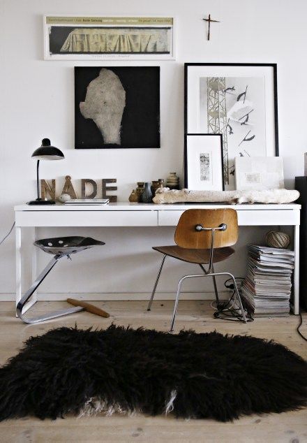 A cozy and artistic corner featuring a white desk with eclectic decor, including a vintage Eames chair, a fluffy dark fur rug on the floor, and an array of interesting art and objects such as a large stone head sculpture, framed abstract artworks, and decorative letters spelling 'NADE'. A desk lamp, stacks of magazines, and assorted pottery complete the curated look.