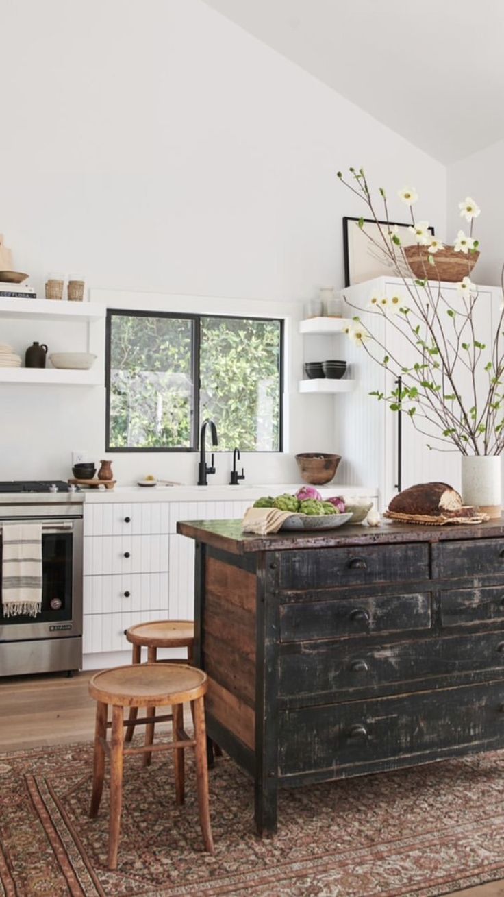 An inviting kitchen featuring a rustic island with a distressed black finish and a wooden countertop, surrounded by a vintage wooden stool. The room has white cabinets with black handles, a built-in oven, and open shelving displaying pottery. A large window allows natural light to illuminate the space, highlighting a bouquet of white flowers in a vase and fresh vegetables on the counter. The warm tones of a traditional patterned rug complement the room's clean, white walls and wooden floor.