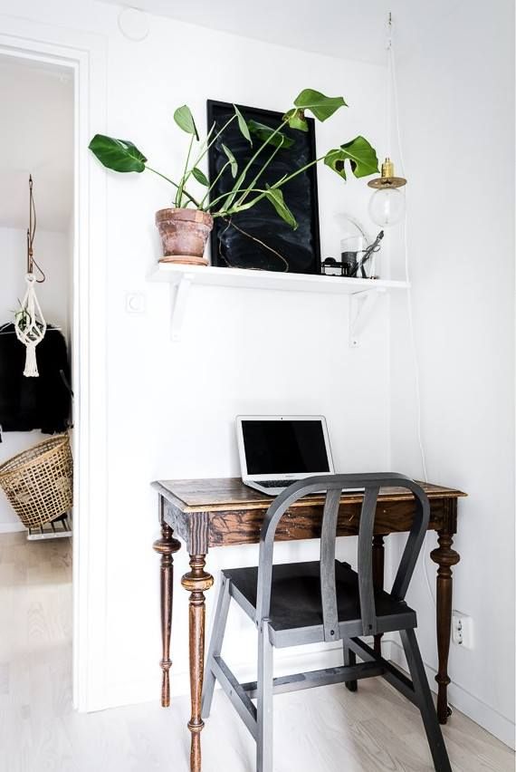 A cozy corner workspace illuminated by natural light, featuring an antique wooden desk with turned legs. On the desk, a laptop awaits use next to a stylish grey bentwood chair. Above, a white floating shelf holds a thriving potted plant with heart-shaped leaves and a simple pendant light, while a woven basket adds a touch of warmth to the black door in the background. The clean, white walls and light wood floors give the space a fresh and airy feel.