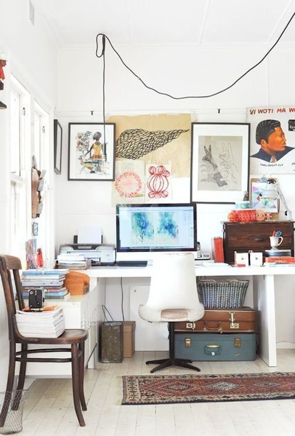 A bright and artistic home office featuring a white desk with a modern computer setup, flanked by stacked suitcases and various printers. The wall above is adorned with diverse artwork including abstract and figurative pieces. A traditional wooden chair and a colorful rug add warmth to the space, complementing the white floors and walls.