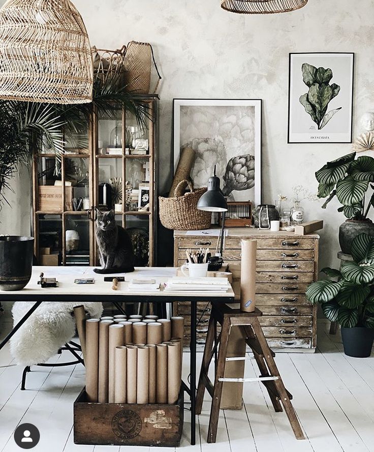 A creative and eclectic workspace featuring a table with rolls of paper, a wooden ladder, a glass-front cabinet filled with various items, lush green plants, and framed botanical prints on the wall, with a cat sitting on the desk.