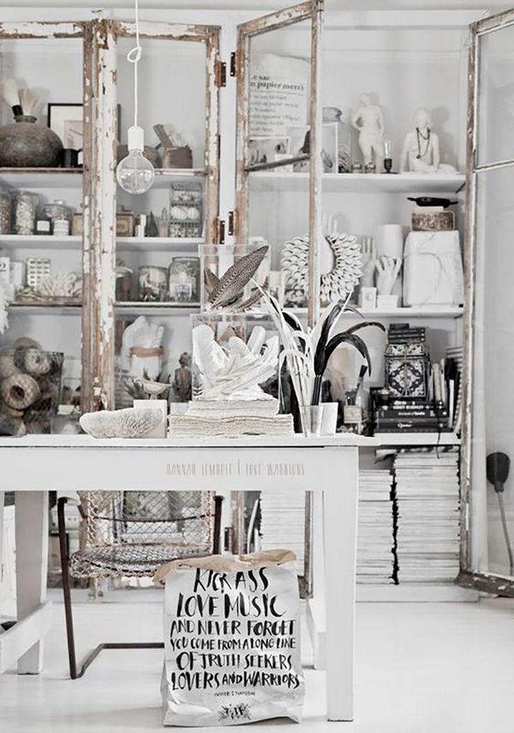 A creative and eclectic workspace featuring whitewashed wooden shelves filled with various decorative items, books, and art supplies. The space includes a rustic wooden table with a whitewashed finish, a collection of feathers and natural elements as centerpieces, and an exposed bulb hanging light fixture, all contributing to a vintage and artistic ambiance.