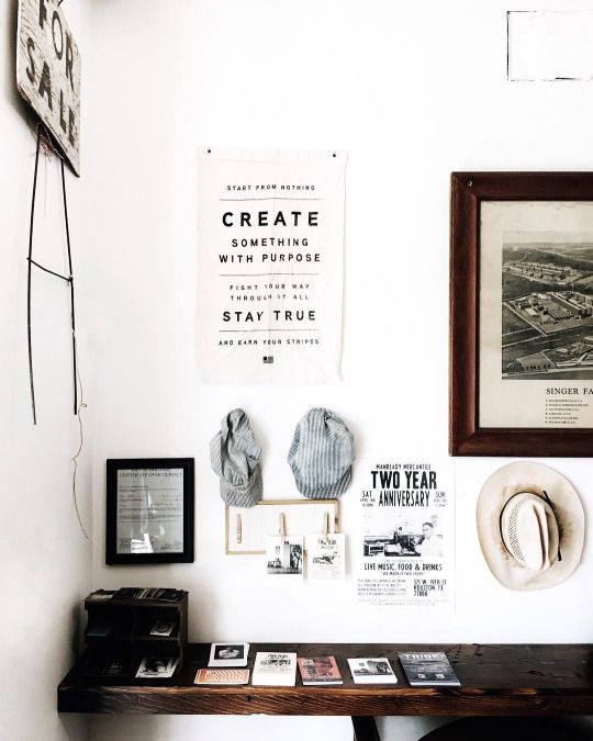 A curated wall display featuring a variety of framed artworks, photographs, and memorabilia. Central to the display is a motivational poster stating "CREATE SOMETHING WITH PURPOSE". Below the arrangement, a wooden shelf holds an assortment of books, a vintage baseball, and other small items.