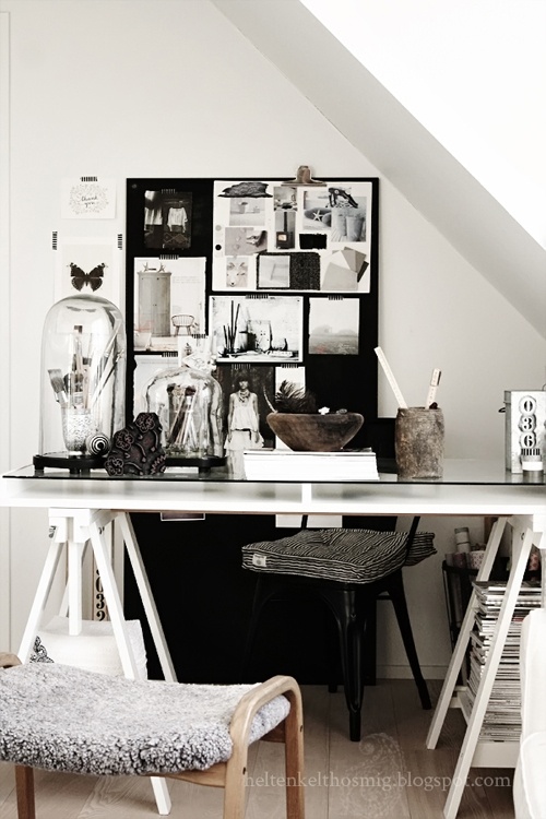A cozy and eclectic workspace set in a small corner, featuring a white sawhorse desk with various decorative items like glass domes, vintage photos on a black pinboard, and rustic wooden bowls. A wooden chair with a textured cushion completes the setup.