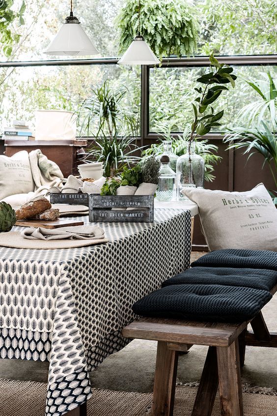 An elegant outdoor dining area featuring a table covered with a patterned tablecloth, surrounded by wooden benches with black cushions. The table is set with decorative items, and lush green plants fill the background, creating a fresh and inviting environment.