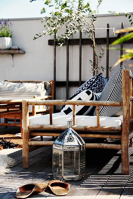 A stylish outdoor seating area featuring a wooden bench with white cushions and black and white patterned pillows. A decorative lantern is placed in front of the bench, and greenery surrounds the space, creating a serene and inviting atmosphere.