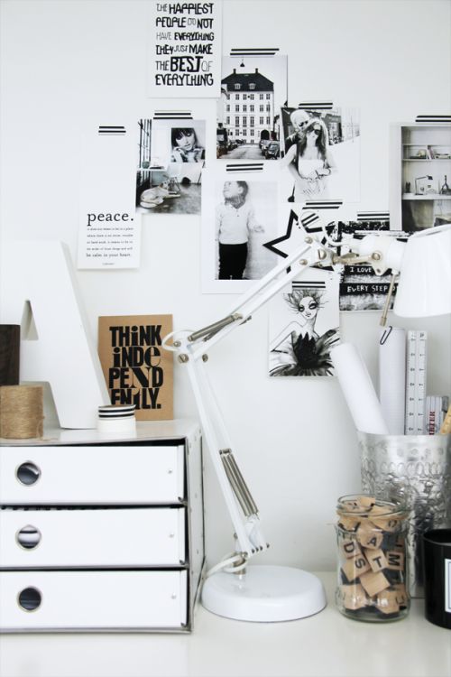 A detailed view of a creative workspace with a focus on an inspiration wall filled with black and white photos, art prints, and motivational quotes. The desk area is neatly organized with a white adjustable desk lamp, a white storage drawer labeled in black, and various office supplies. A glass jar holds a collection of scrabble tiles, adding a personal touch to the decor. The overall ambiance is modern and inspirational.