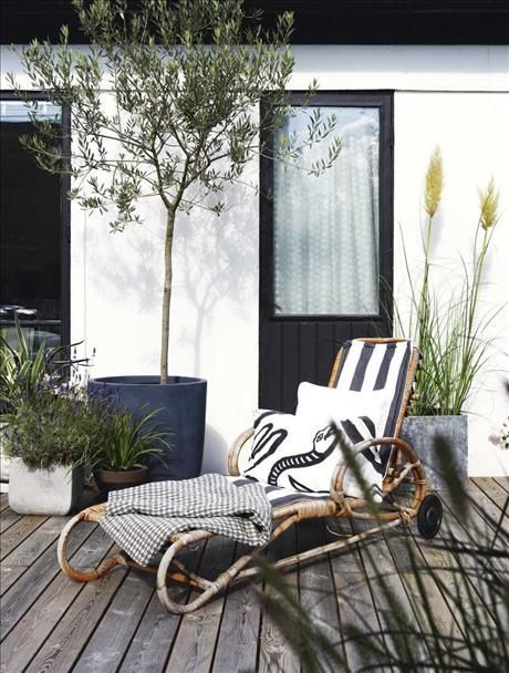 A stylish outdoor deck featuring a rattan lounger with a striped black and white cushion and a matching pillow. The space is enhanced by potted plants, including a small tree and various green foliage, set against a modern white and black exterior.