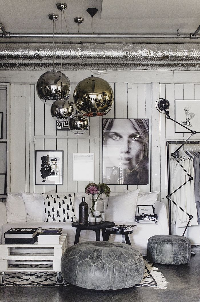 A stylish living room with white walls and industrial decor. The room features a white sofa with patterned cushions, a rustic wooden pallet coffee table, a large round gray ottoman, and metallic spherical pendant lights hanging from the ceiling. Black and white artwork, including a large portrait of a woman’s face, adorns the walls.