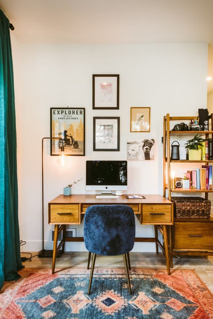 A cozy home office featuring a mid-century modern desk with a desktop computer, surrounded by a variety of framed artworks on the wall and a wooden bookshelf filled with books and items. A vibrant multicolored rug lies on the floor, complementing the emerald green curtains.
