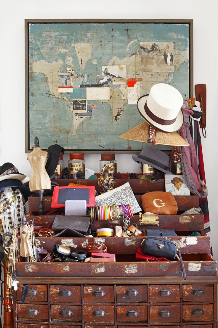 This image features a vintage, multilayered display of diverse objects arrayed on an aged wooden apothecary chest. Above the chest hangs a large, weathered world map framed in dark wood. The chest drawers are open, revealing an eclectic mix of items including hats, scarves, a miniature mannequin, colorful bangles, old books, and various bags. The assortment conveys a rich sense of travel and collection over time, blending fashion elements with a global aesthetic. The overall feel is that of a personal treasure trove, showcasing a life of adventures and discoveries.