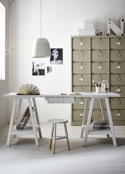 A minimalist home office with a white sawhorse desk, a small stool, and an industrial metal filing cabinet. The workspace is decorated with a large white pendant light, monochrome photos, and simple stationery items.