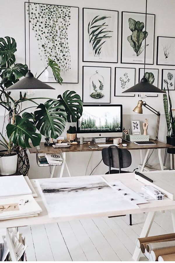 An artistically arranged home office featuring a collection of botanical prints on the wall. The prints, which include various plant species, are elegantly framed and hung above a workspace that includes a desk with a desktop computer, surrounded by live plants like monstera and smaller potted greens. The room has a minimalist vibe with white walls and a light wooden floor, accentuated by two industrial-style pendant lights over the desk. Art supplies and personal items are neatly organized on the desk, creating a functional yet stylish creative space.