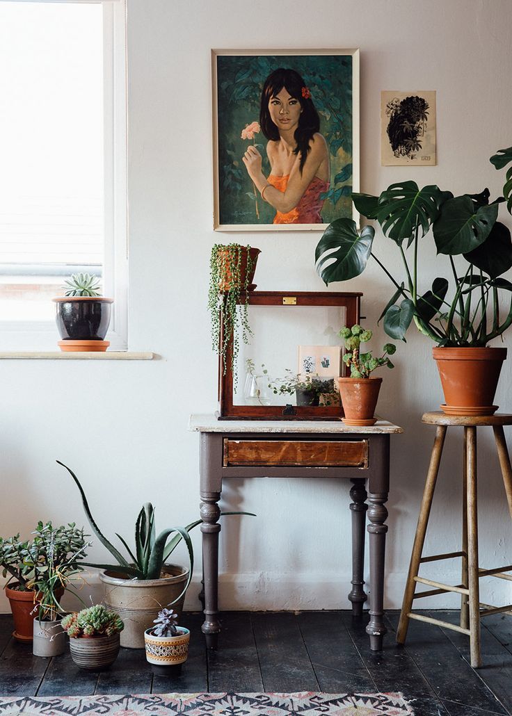 A cozy corner with a vintage wooden table displaying an array of potted plants and a large portrait of a woman holding a flower. The space is complemented by additional artworks, a vintage mirror, and plants on varying levels, creating a layered, textured look typical of boho interiors. The dark wooden floor and rug add warmth to the scene.
