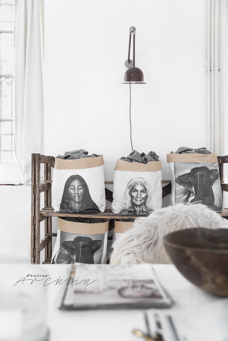 A minimalist loft space with white walls and wooden accents. The room features a set of white laundry baskets with black and white portraits printed on them, a rustic wooden shelf, and an industrial-style wall lamp. The space is decorated with simple, monochromatic elements creating a clean, modern look.