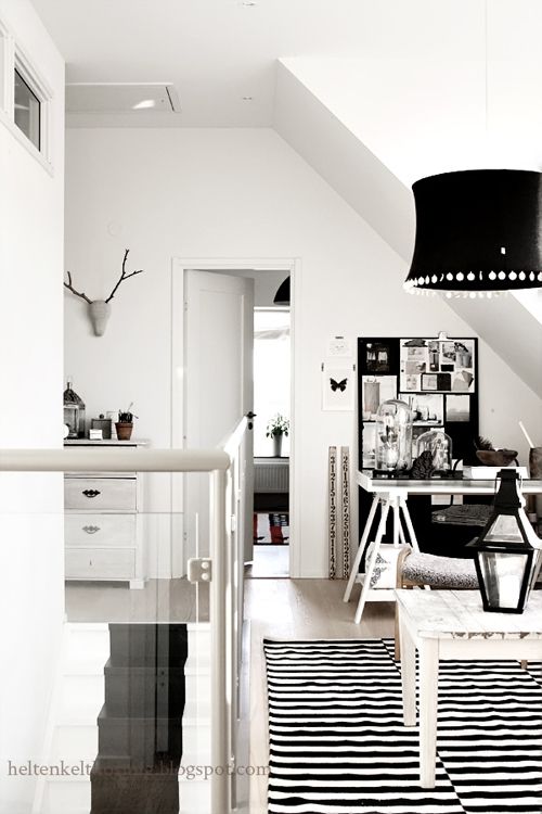 A chic attic workspace featuring white walls and a sleek white desk with an inspiration board filled with notes and images. The area includes a striped black and white rug on the floor and a decorative hanging lamp with fringes. Near the desk, a small white drawer unit and various decor items like lanterns and jars add to the aesthetic. The space is accented with a minimalist antler decor on the wall.
