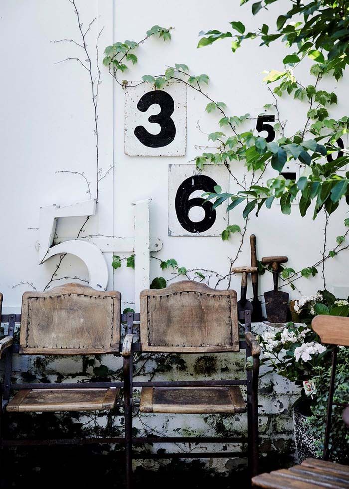 An outdoor seating area with vintage wooden chairs against a white wall decorated with large black and white numeric signs. The area is surrounded by green plants and climbing vines, creating a rustic and charming atmosphere.