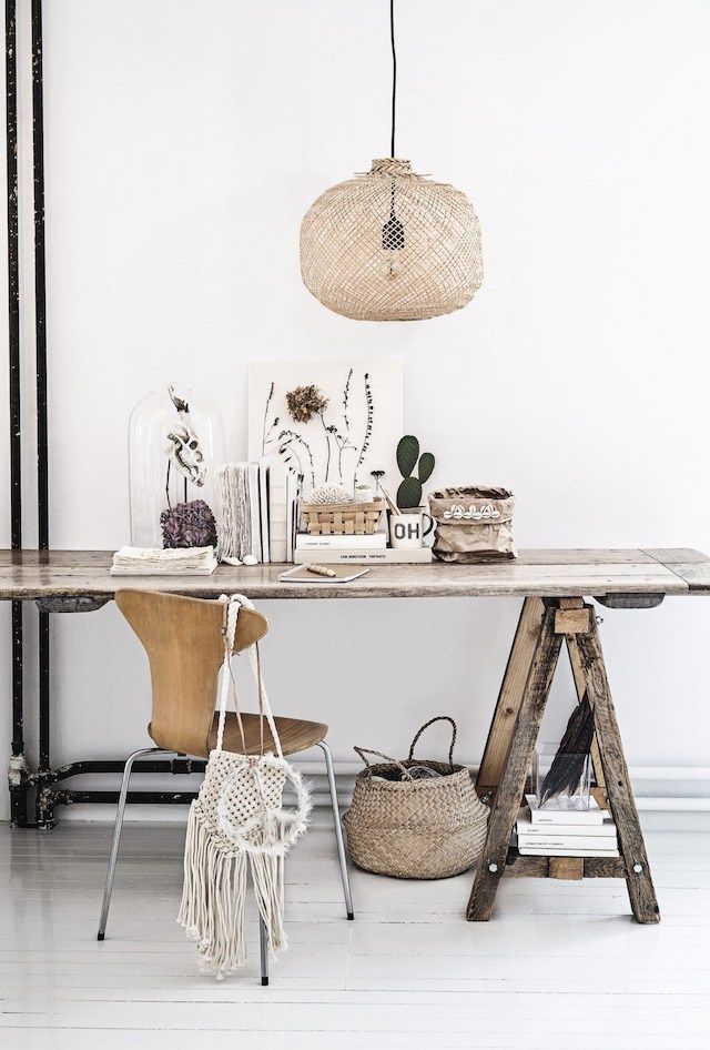 A Boho-style workspace featuring a rustic wooden table with a variety of decorative items including glass domes, dried flowers, books, and a cactus. A wicker pendant light hangs above, and a woven bag is draped over a wooden chair.