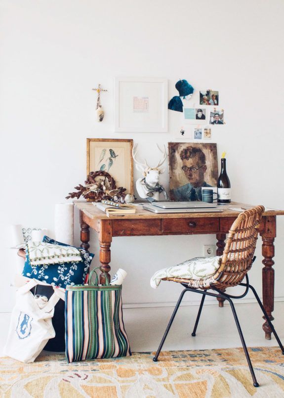 An eclectic artist's workspace featuring an antique wooden desk cluttered with various art pieces, a painting of a man, and multiple eclectic objects. A wicker chair with a decorative throw sits next to the desk, and an artistic tote bag leans against it, set against a neutral wall and on a patterned light rug.