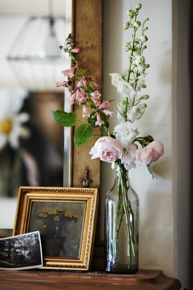 A serene vignette of floral arrangements in glass bottles and vases, framed botanical prints, and art books on a white console table.