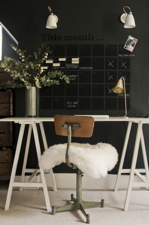 A modern and minimalist home office setup featuring a white trestle desk on a plush white rug. The wall behind the desk is painted in a deep charcoal and serves as a chalkboard calendar labeled "This month," filled with handwritten notes and reminders in white chalk. A rustic industrial chair with a green metal frame and a worn wooden seat is covered with a fluffy white sheepskin throw, adding a touch of comfort. To the right of the desk, a small gold clip lamp provides task lighting. The scene is further enhanced by a tall vase filled with eucalyptus branches, placed on the desk, lending a refreshing touch to the space.