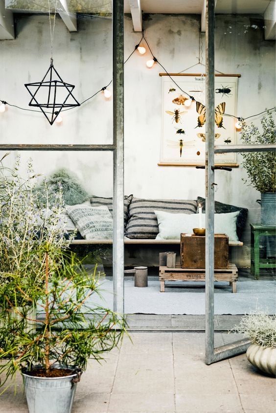 A cozy indoor seating area with a rustic bench adorned with striped and patterned pillows. The space is decorated with green plants, string lights, and a hanging geometric ornament, all visible through large glass doors.