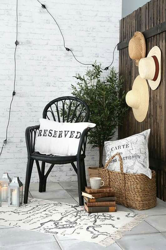 A cozy corner of a patio with a black wicker chair adorned with a white pillow that reads ‘Reserved.’ The area is decorated with hanging hats, a potted plant, a wicker basket, string lights, and a rug, creating a charming and personalized outdoor space.