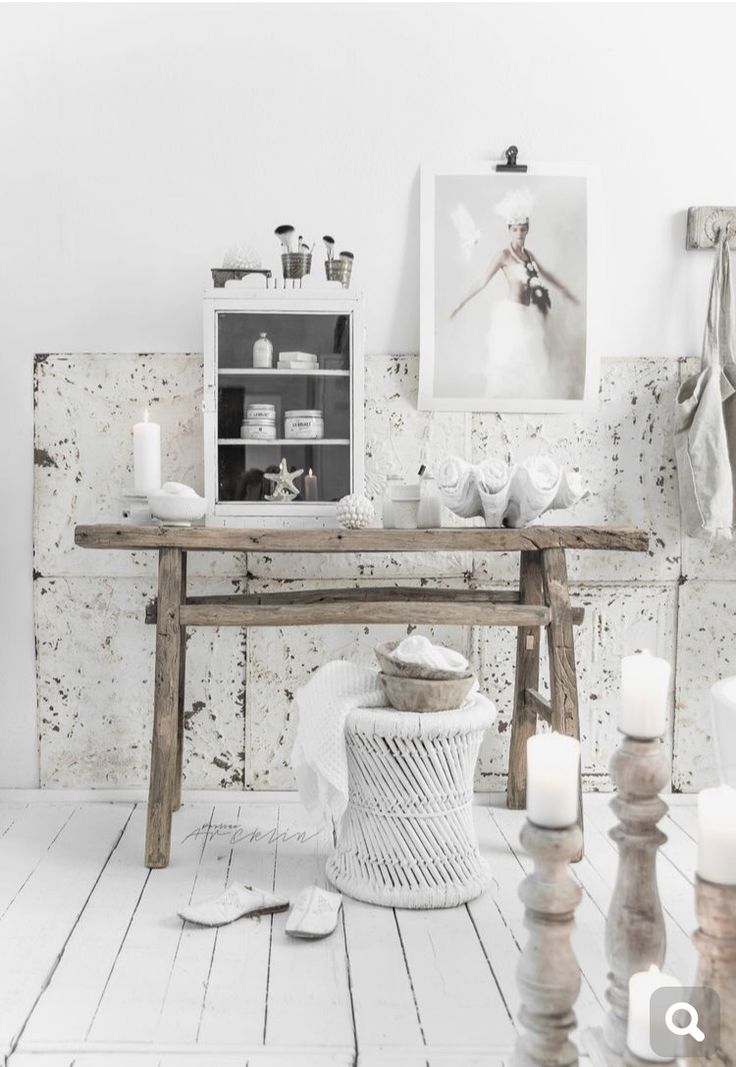 A serene and rustic workspace with whitewashed walls and a wooden table. The decor includes a white wicker stool, tall white candles on wooden holders, and a large black and white photograph of a ballerina on the wall. The space is bright and airy, with an emphasis on natural and minimalist elements.