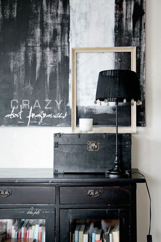 A chic and stylish vignette showcasing a black cabinet with glass doors filled with books, topped with a black lamp and a framed piece of abstract art. The backdrop features a large black and white abstract painting with the words ‘Crazy about fragrances’ written in elegant script, creating a modern and sophisticated atmosphere.