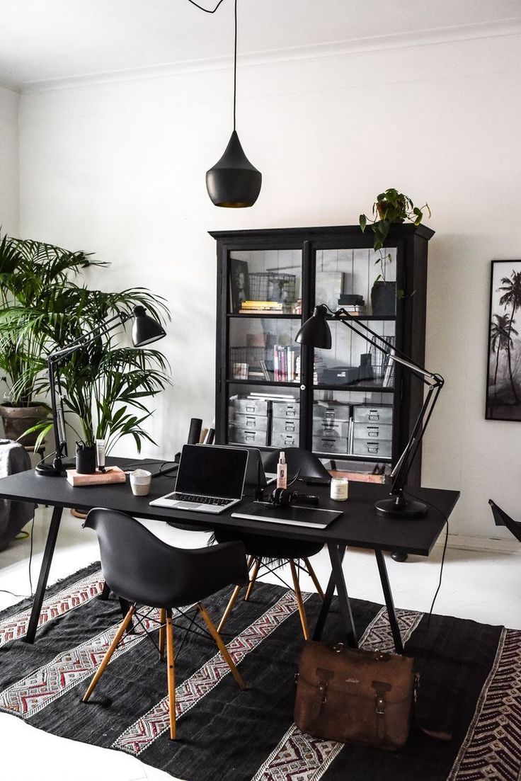 A stylish and modern home office setup with a minimalist black desk, equipped with two laptops, a tablet, and an adjustable desk lamp. There are two Eames-style black chairs with wooden legs, and a sleek, dark cabinet filled with books and decor items. The room is decorated with several indoor plants and features a geometric-patterned rug. A hanging black pendant light adds a touch of sophistication to the space.