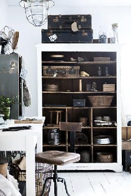 A stylish, rustic room with a large wooden shelving unit filled with vintage items and storage baskets. The shelves contain various objects, including suitcases, jars, and woven baskets. The space also features a vintage office chair, a white desk with a metal wire basket underneath, and industrial-style lighting, creating a charming and functional workspace.