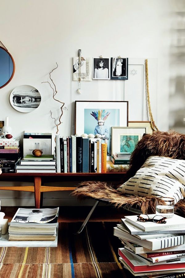A cozy living space with a wooden bench filled with books and framed artwork. A furry throw and patterned pillow adorn a nearby chair, and a stack of books on the floor adds to the layered, bohemian vibe. The wall features a mix of mirrors, photos, and hanging decor.