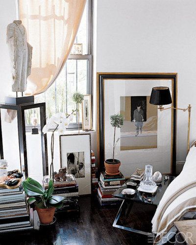 A serene bedroom corner with a mix of stacked books, framed art, and potted plants. A small table holds a lamp and decorative items, while natural light filters through sheer curtains, creating a calm and inviting atmosphere.
