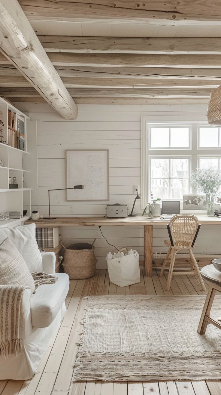 A cozy, light-filled cabin interior with a white couch, wooden ceiling beams, and a large window allowing natural light to fill the space. The decor includes a wooden desk with a wicker chair, a light beige rug, and various neutral-toned accessories.