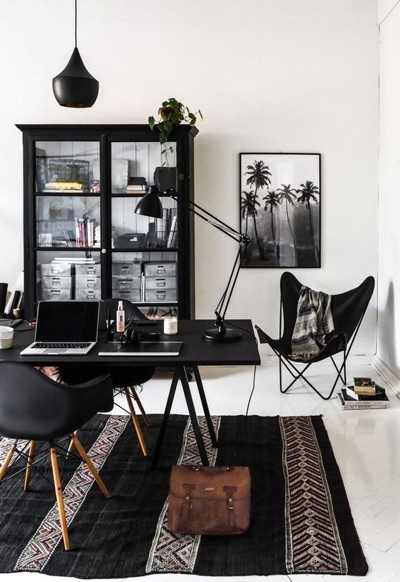 Masculine home office with black and white color scheme, featuring a black desk, black chair, black cabinet with glass doors, a black pendant light, and a framed black and white photo of palm trees. The office is decorated with a dark patterned rug, a leather bag, and plants.