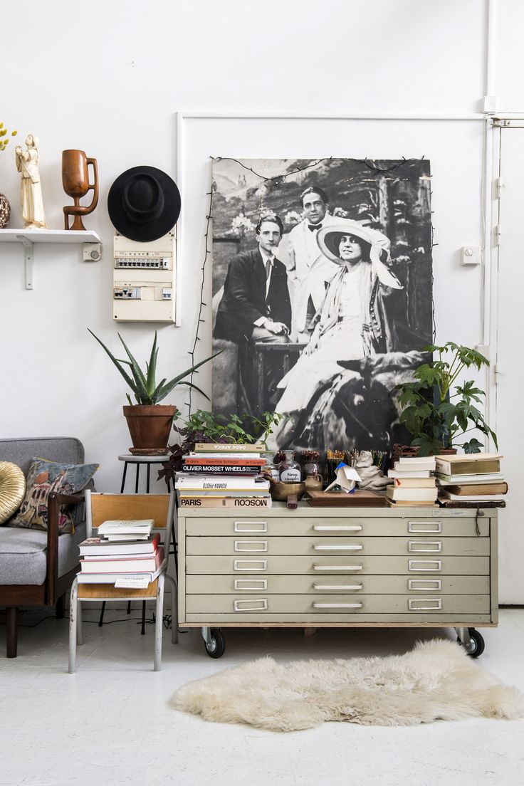 A stylish living space featuring a large vintage family portrait hanging on a white wall. The decor includes a mix of modern and vintage elements such as potted plants, books, and eclectic accessories. The portrait is black and white, showing three people from the early 20th century.