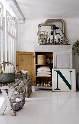 A bright and airy room featuring a vintage wooden cabinet with open shelves displaying neatly folded linens, jars, and decorative items. A large green letter “N” sign leans against the cabinet. The room also includes a rustic bench with a metal basket underneath, and various decor pieces such as lamps, mirrors, and vases, creating a harmonious and inviting atmosphere.